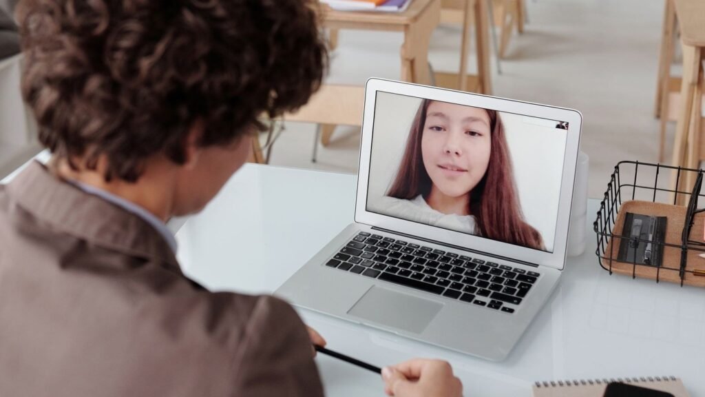 Docente de espaldas dando una clase virtual a una alumna que aparece en la pantalla de una notebook